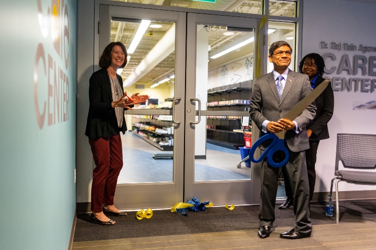 Chancellor Agrawal holds a pair of giant scissors in front of the doors of the Care Center. Ribbon pieces lay on the floor and his wife, Sue, is smiling and applauding.