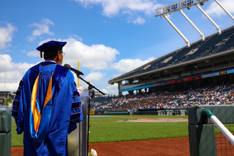 Chancellor Agrawal stands at a podium on the field of Kauffman Stadium with the stands in the background