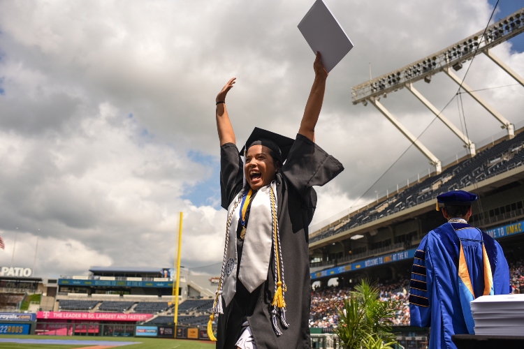 A graduate stands on the stage with her hands in the air, diploma in one hand, smiling
