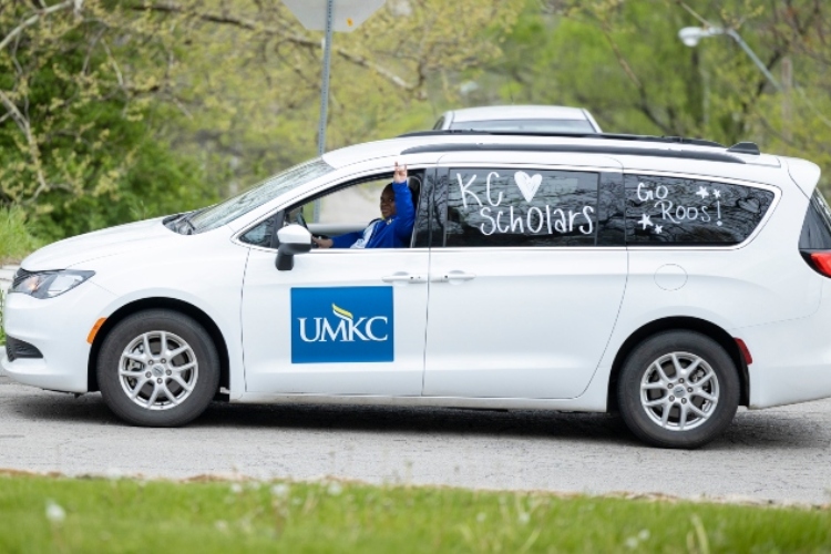 A person sits in a white car with a UMKC logo on the driver door and does a Roo Up hand gesture out of the window. The back driver-side windows have window painting that say "KC Scholars" and "Go Roos"