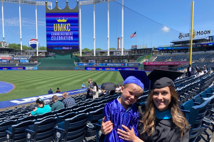 A mother and son in graduation robes pose for a photo in front of Crown Vision at Kauffman Stadium