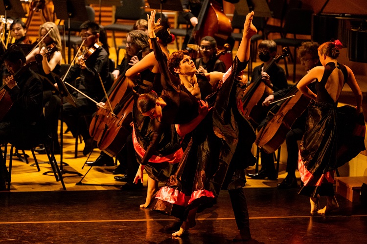Two dancers perform in front of a string ensemble on a lit stage