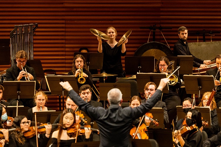 A conductor has his back toward the camera; in front of him is an orchestra performing onstage.