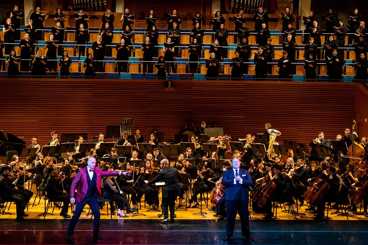 Two opera singers perform in front of an orchestra on a lit stage