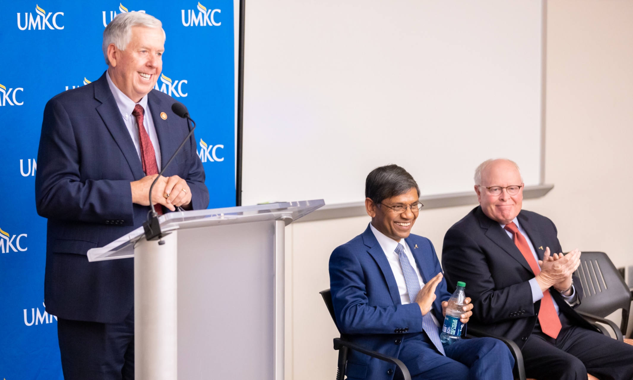 Governor Mike Parson stands smiling at a podium with a UMKC backdrop behind him. Chancellor Mauli Agrawal is seated to the governor's left, smiling.