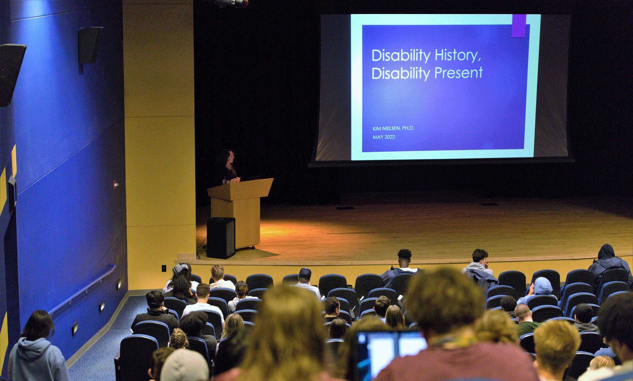 Kim Nielsen stands on a stage behind a podium, with a large screen reading, "Disability History, Disability Today" to the right of her. The photo is taken from partway up theatre seating, so the backs of many students heads are visible between the camera and Nielsen.