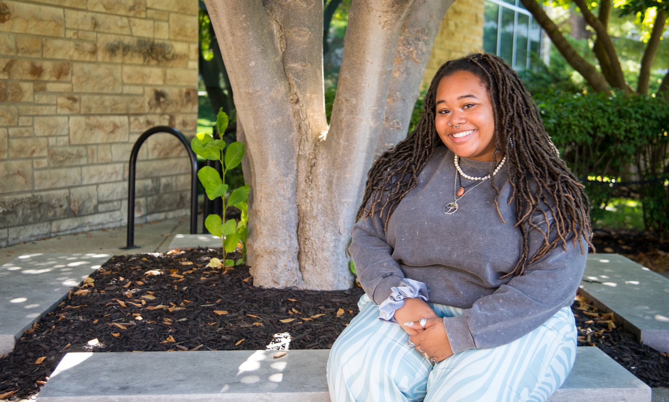 Ophelia Griffin, wearing jeans and a gray sweater, sits on a bench in front of a tree and smiles at the camera.