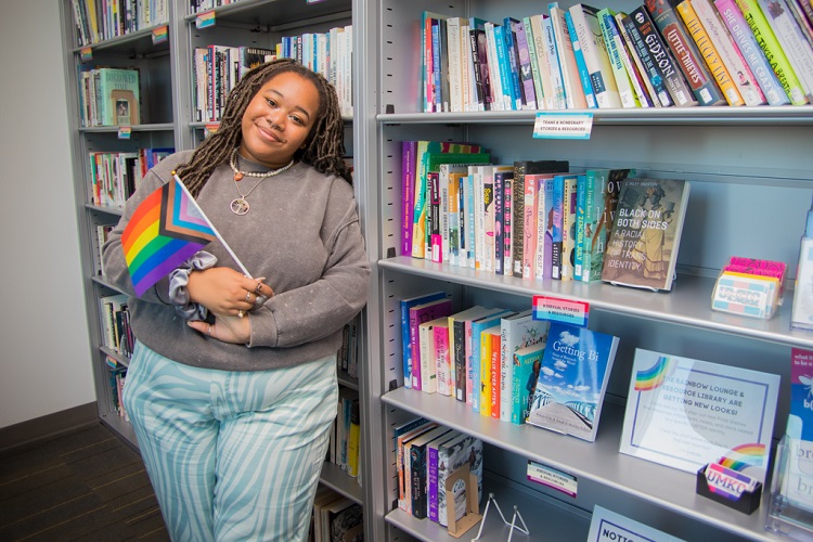 Ophelia Griffin stands next to a bookshelf in the Rainbow Lounge, filled with books to the ceiling. She holds a progress pride flag in her hands with her arms at her torso in front of her.