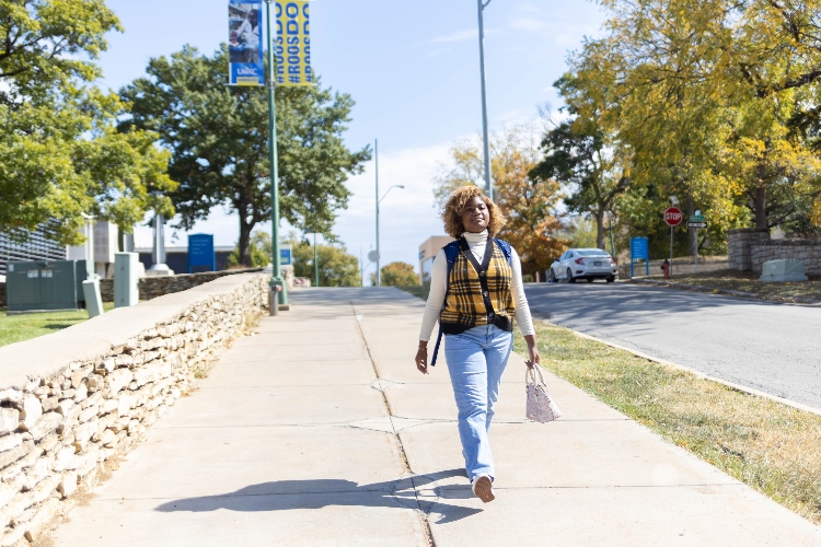 Jayla Williams walks down the sidewalk on the UMKC campus with her backpack and purse. In the background you can see UMKC signs.