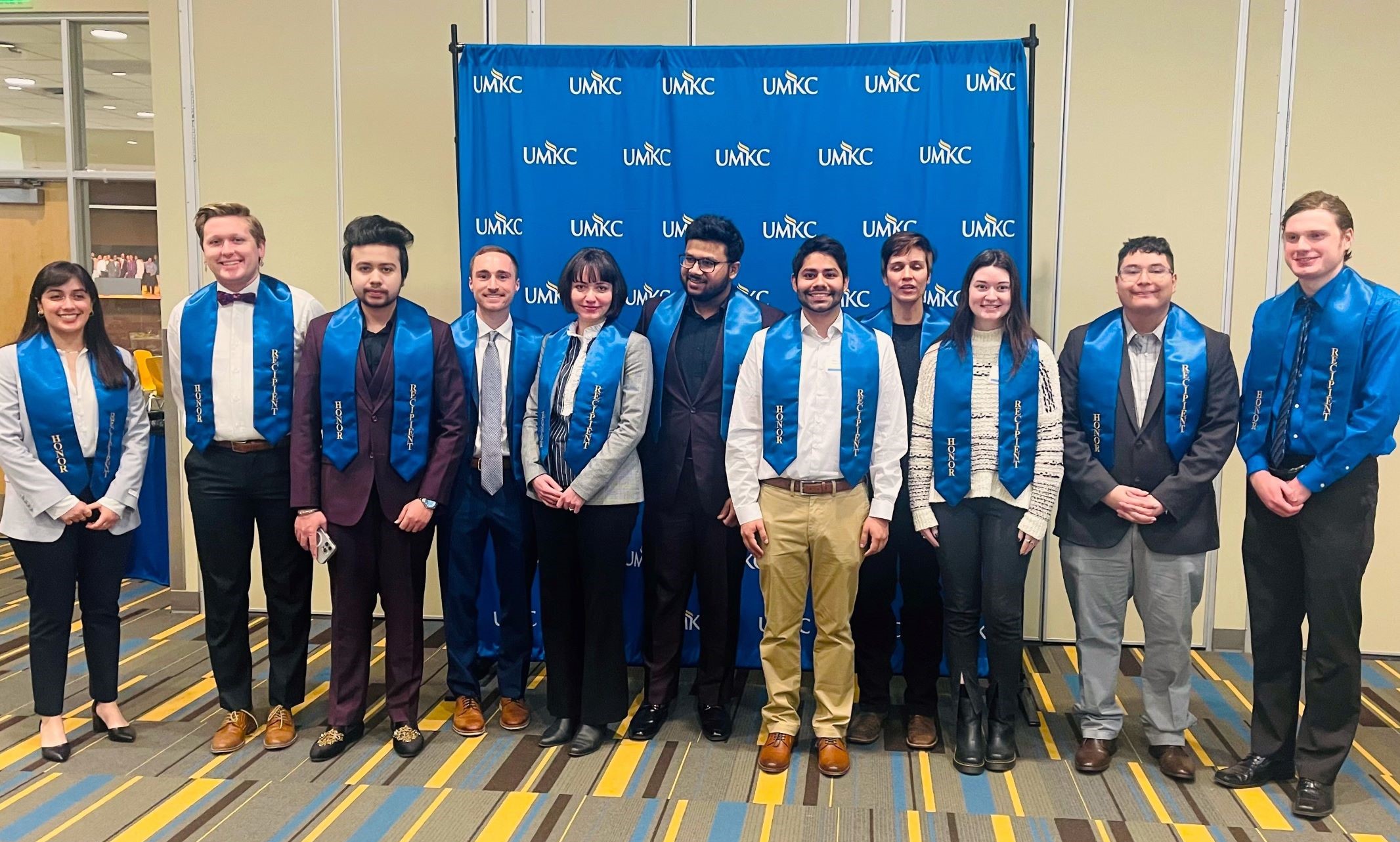 Fifteen students pose for a photo in front of a UMKC backdrop