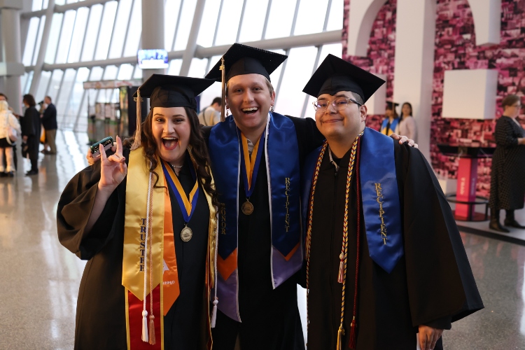Three graduates smile and one holds a Roo Up inside the rotunda of the T-Mobile Center