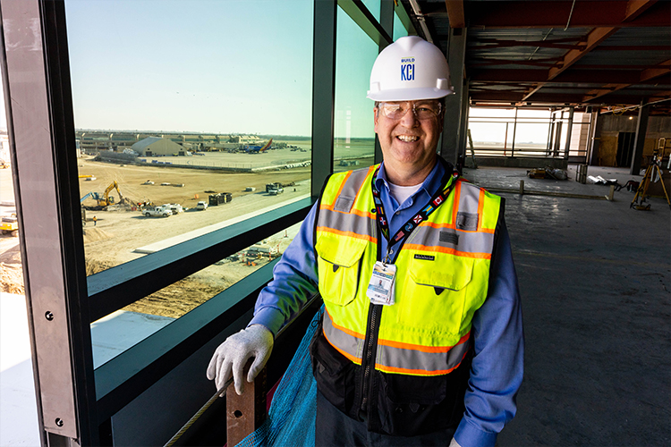 Joe McBride stands in construction gear and smiles at the camera. The old airport is behind him.