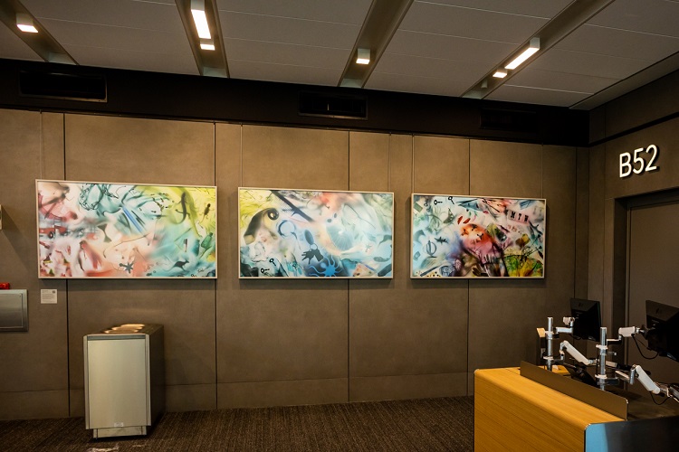 Kati Toivanen's art on the wall by Gate B52. The artwork is three horizontal panels of colorful, dreamy-looking items in an "I Spy" fashion.