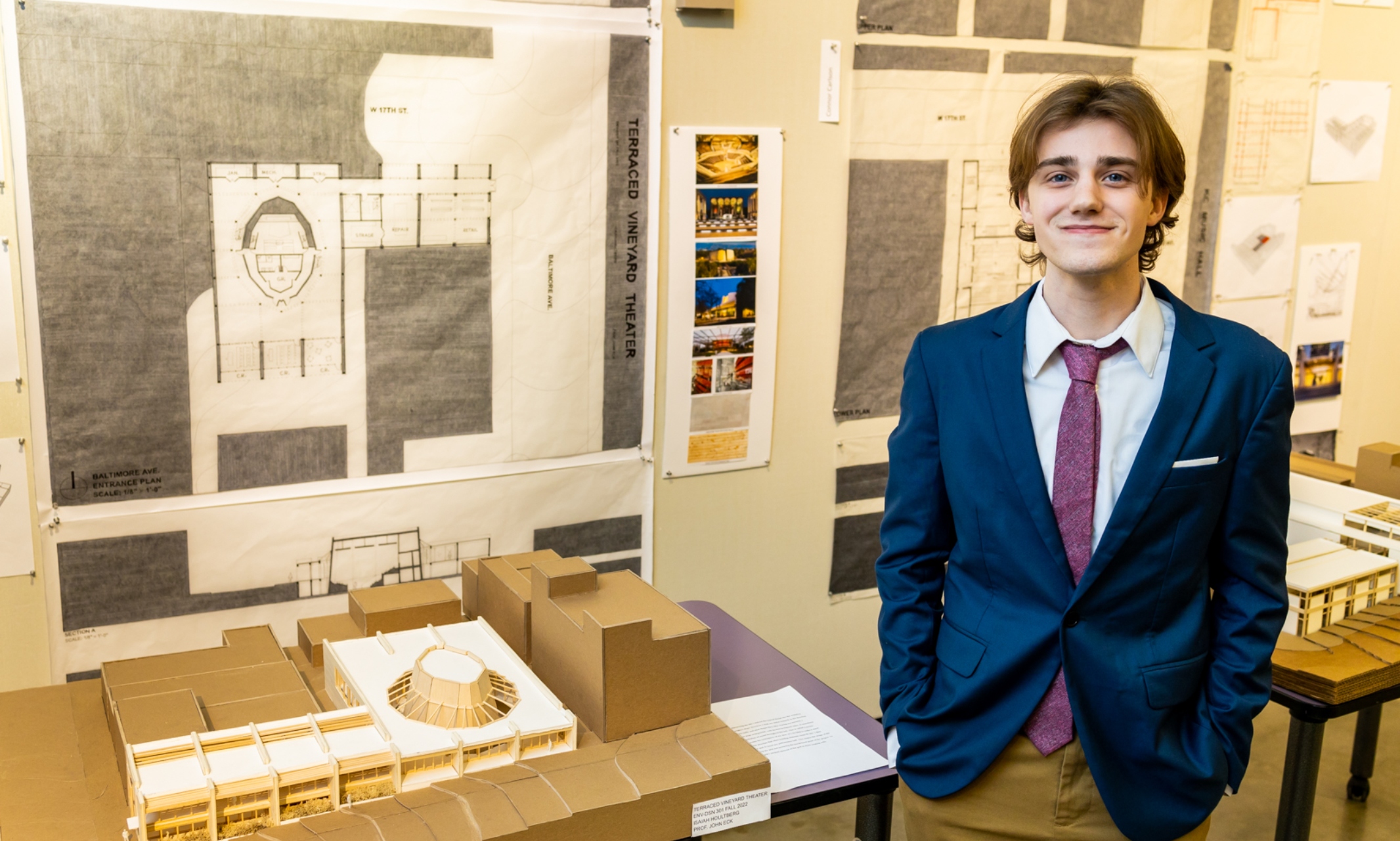 Isaiah Houltberg stands with his hands in his pockets, smiling next to his design model