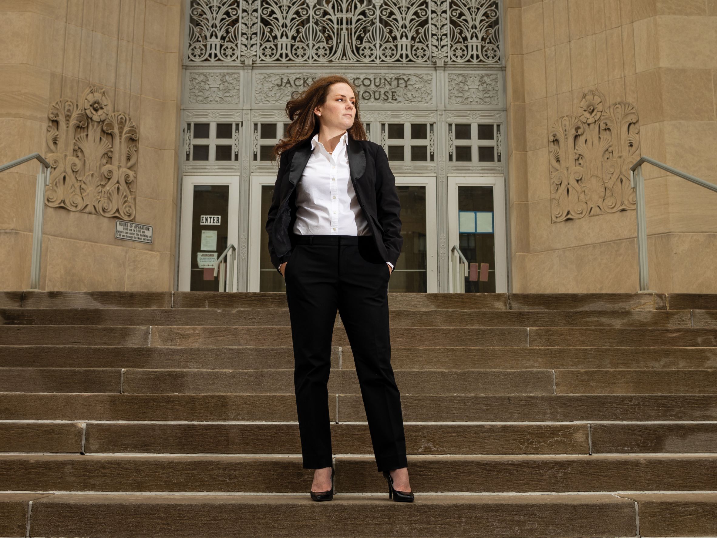 Audrey McCormick stands on the steps of the Jackson County Courthouse in a powerful pose with her hands in her pocket. She is looking off to the side with a serious expression. The wind is blowing her hair