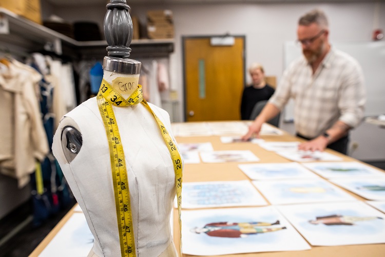 A mannequin with a measuring tape is in the forefront. Behind it is a large table with several ilustrations on it. A professor and student look at the illustrations.