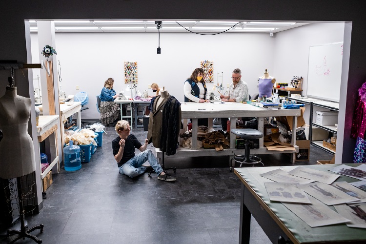 Students and a professor work in a studio. One sews, two look at illustrations on a table and another looks at costumes.