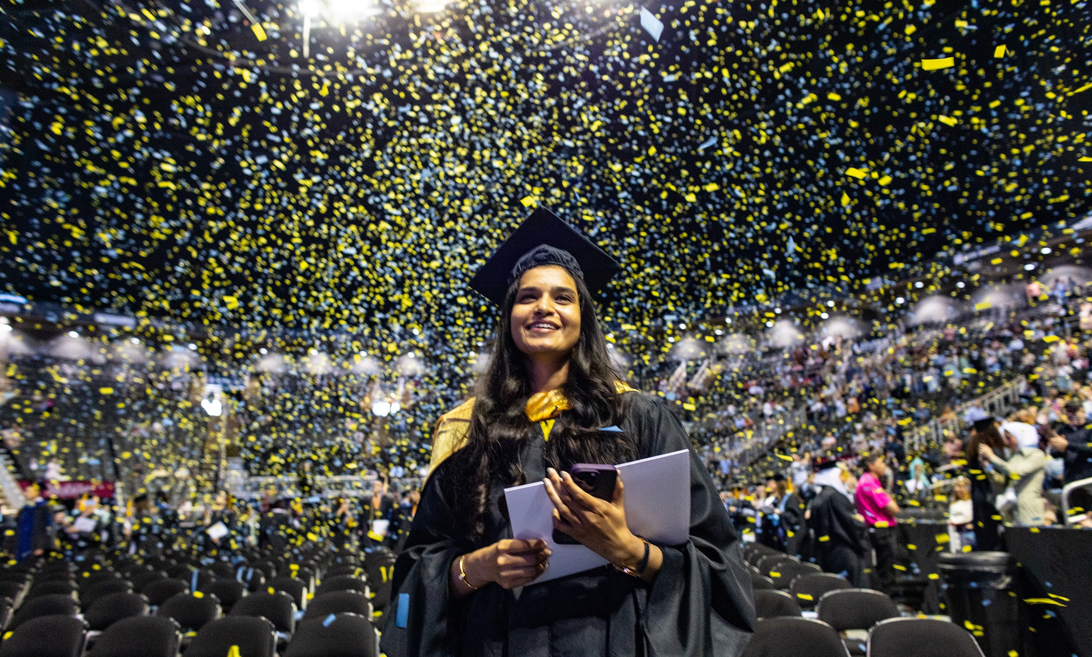 Graduating student stands facing the camera and smiling. Blue and gold confetti rains down behind her and other graduates