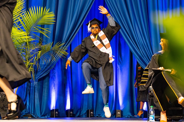 A student is mid-jump as he dances on stage while receiving his diploma