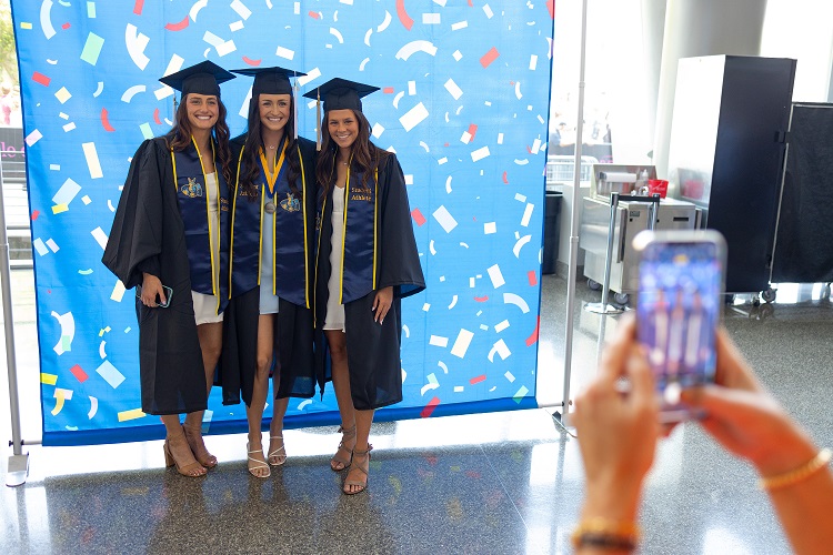 Three graduating students pose together in front of a backdrop as someone takes a photo of them on a phone