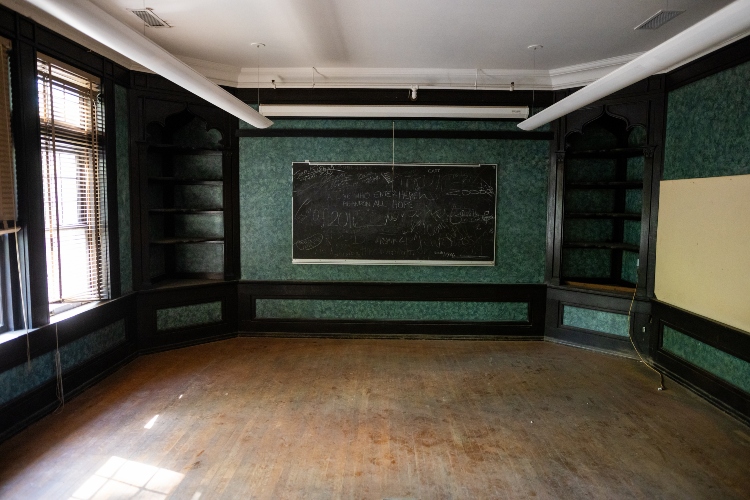 A wide shot of a room with a black chalkboard across the back wall. In the corners there are built in black bookshelves. An ornate green wall paper lines the walls