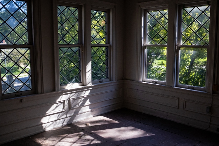 A corner of a room lined with large windows on each side. The sun is beaming in and there are lush trees outside the window.