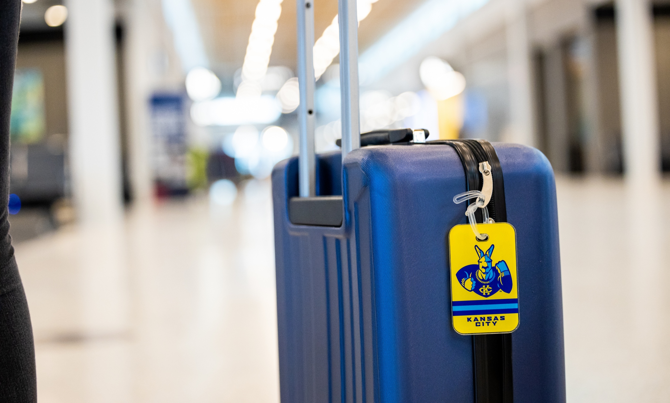 A close up of a blue suitcase with a yellow UMKC luggage tag that has a Roo on it with the KCI airport terminal in the background