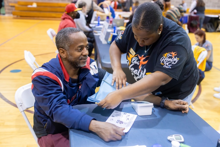 A person helps another person with a blood pressure cuff at a health event inside a gymnasium filled with people