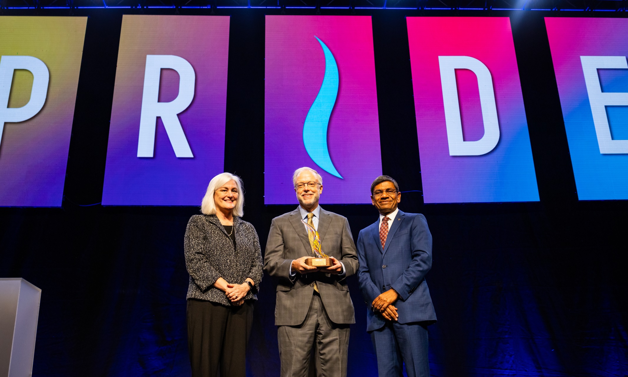 Madeline McDonough, Don Hall Jr. and Chancellor Mauli Agrawal on stage. The large screen behind them reads "PRIDE" in colorful letters.