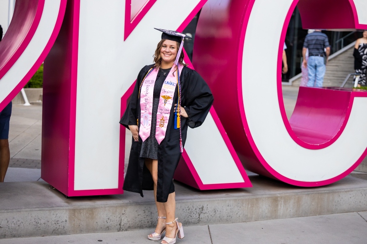 Macila Arnold stands in front of a large, bright pink KC sign smiling in a cap and gown