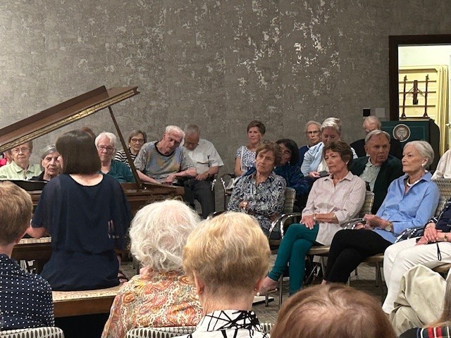 Beth plays the piano, surrounded by an audience of Claridge Court residents sitting in chairs and listening
