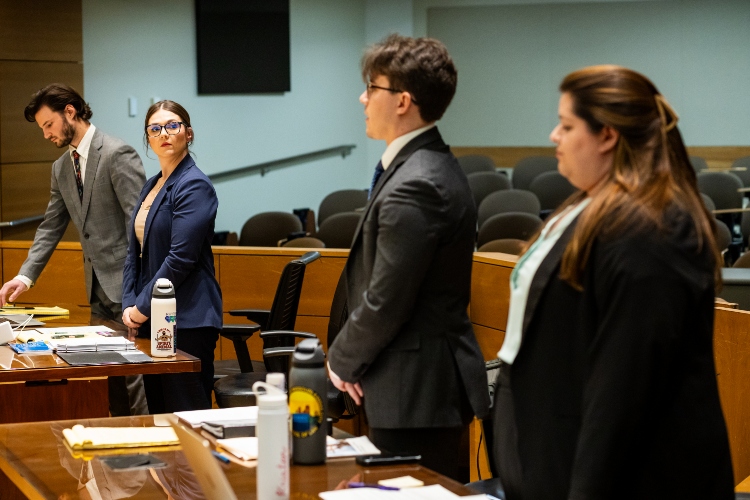 Students stand in a mock courtroom in business attire at the counsel benches
