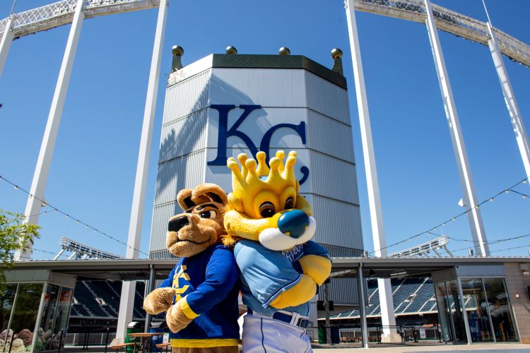 KC Roo and Sluggerrr pose standing back-to-back in front of the CrownVision board at Kauffman Stadium