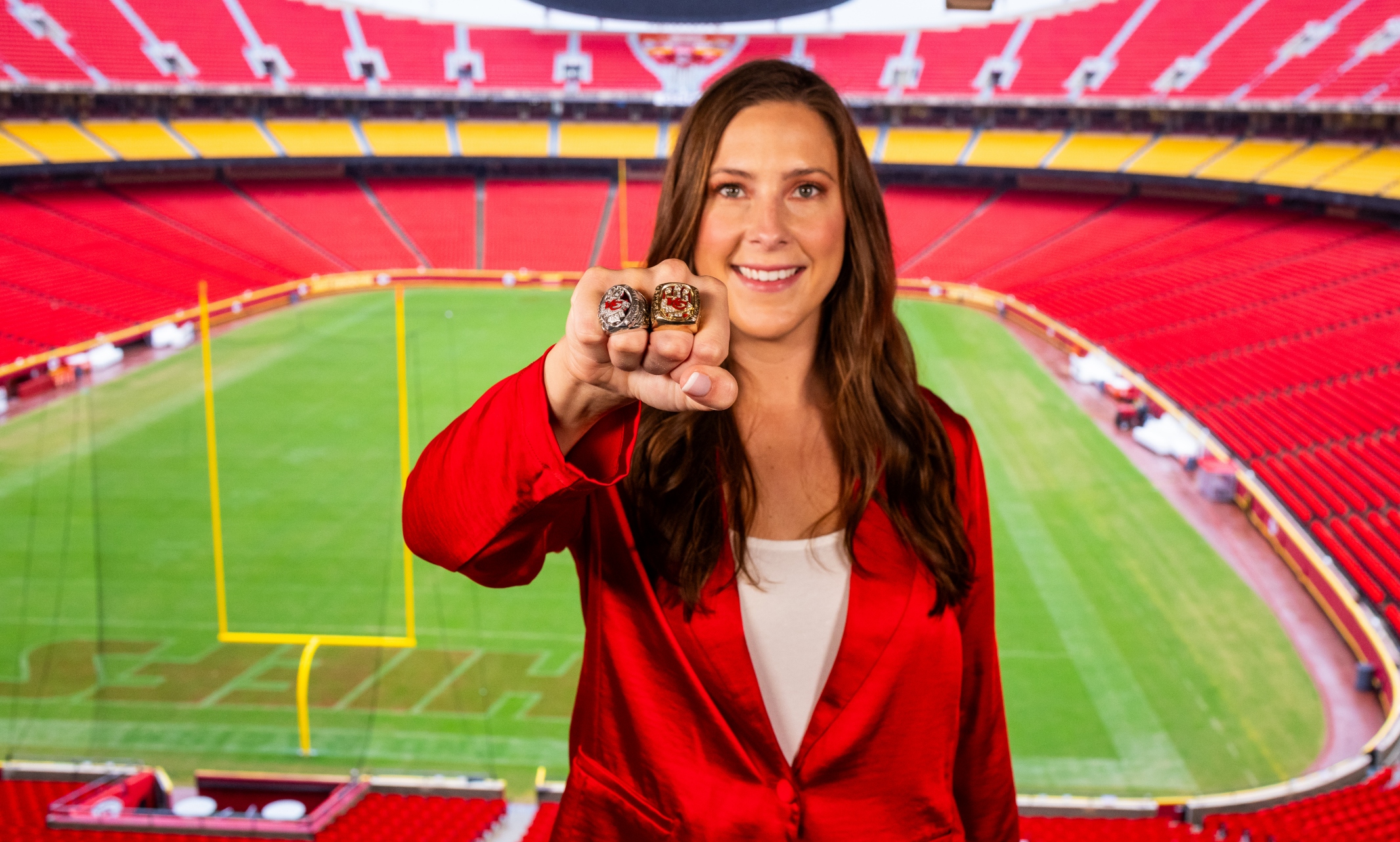 Meghan Jaben stands in the seating bowl of Arrowhead Stadium overlooking the field. Standing in a red blazer and smiling, Meghan's arm is outstretched in front of her in a fist with two super bowl rings on her fingers.