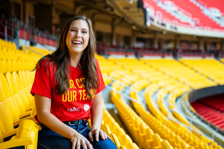 Meghan Jaben smiles seated in the yellow club seats at Arrowhead with a red shirt that says "Our City, Our Team" and the UMKC and Chiefs logos