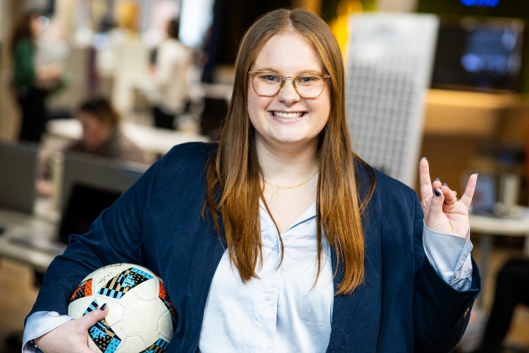 Annmarie Orlando holds a soccer ball under one arm and a roo up gesture with the other hand inside the Sporting KC office