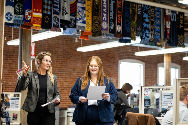 Annmarie Orlando and her mentor Kylie DeWees walk through the Sporting KC office together with rows of soccer scarves hanging from the ceiling rafters above them
