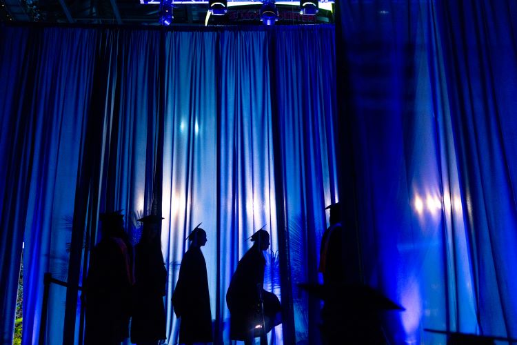 Graduates are silhouetted against blue curtains, waiting to walk across the stage