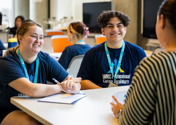 Two people in dark blue shirts with lanyards talk to someone not facing camera