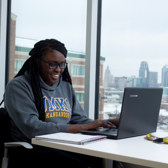 A student smiling at her computer as she takes an online learning course while sitting in front of an apartment window that overlooks the city. 