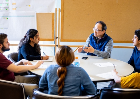 A professor animatedly explaining course concepts to students while sitting around a large table.