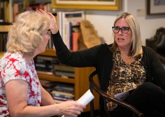 Two people sit talking to each other. The person whose face is in frame has blonde shoulder-length hair and is wearing a printed shirt with a black sweater and is using their hand to talk.