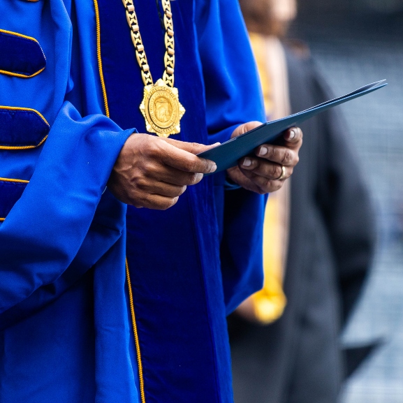 A closeup view of a faculty member wearing a blue and graduation robe and medallion while holding a folder.