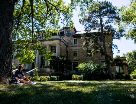 Student wearing blue shirt sits in grass by a tree outside of a building on campus. The student has shoulder length hair and has a laptop on their lap.  