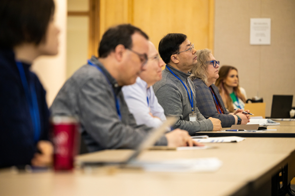 Several faculty members wearing suits sit in a row of a lecture hall looking at lecturer out of frame. 