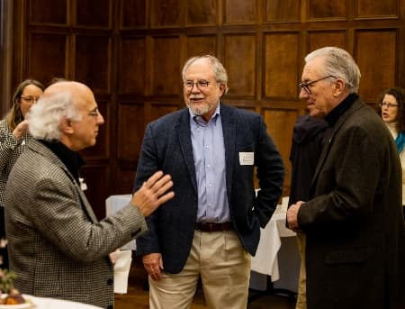 Faculty stand in a group talking. One person is smiling wearing a green shirt