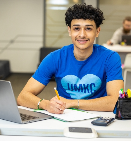 student wearing a blue UMKC shirt with a heart stares at the camera and holds a pencil while sitting at a table. The table has a laptop and folder with paper on top of it.