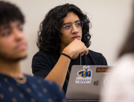 A student sitting in class listening intently to his instructor while taking notes on a laptop. 