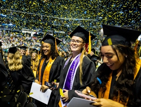 A group of students dressed in their caps and gowns smiling and celebrating during the commencement ceremony as confetti rains down from above.