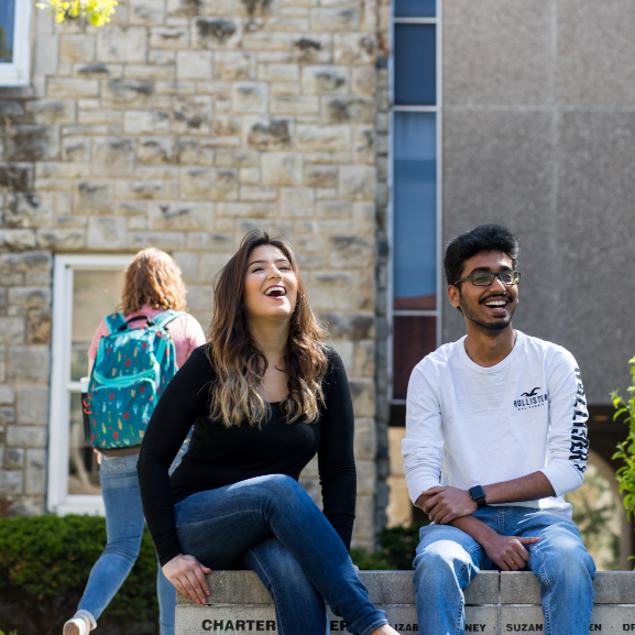 Two students talking and laughing while sitting on a wall on campus.